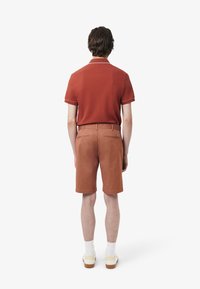Polo corto arancione con rifiniture bianche, abbinato a pantaloni corti arancioni. Tasche posteriori visibili; il modello indossa calzini bianchi e scarpe chiare.