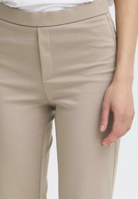 Pantaloni beige su misura con una texture liscia, caratterizzati da una linea dritta, tasca frontale e una vita lucida. Mano posizionata vicino al tessuto.