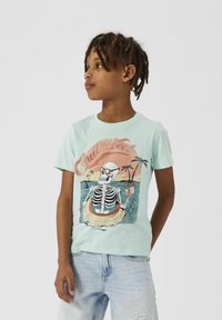 Camiseta verde menta con un gráfico de un esqueleto con gafas de sol en un salvavidas, con palmeras y un fondo de atardecer.