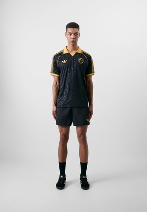 Zwarte sporttrui met een gele kraag en accenten, voorzien van een subtiele tonale patroon. Gecombineerd met zwarte shorts en zwarte sneakers.