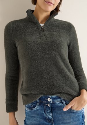 Kvinde iført mørkegrå fuzzy quarter-zip sweater og blå jeans med hånden i lommen, stående mod en ensfarvet væg.
