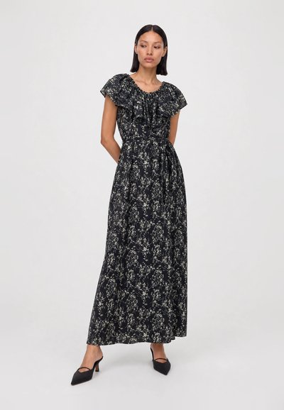 Woman standing wearing a black floral maxi dress with ruffled neckline and short sleeves, paired with black pointed heels.