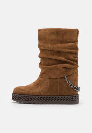 Botte mi-mollet en daim marron avec tige ample, semelle épaisse avec détail chaîne et une chaîne métallique décorative enroulée autour du talon.
