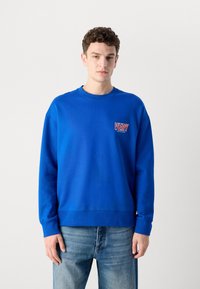 Moletom azul com gola redonda em canelado, ombros caídos e um logótipo frontal que diz "VERY" em vermelho e branco. Usado com jeans azul claro.