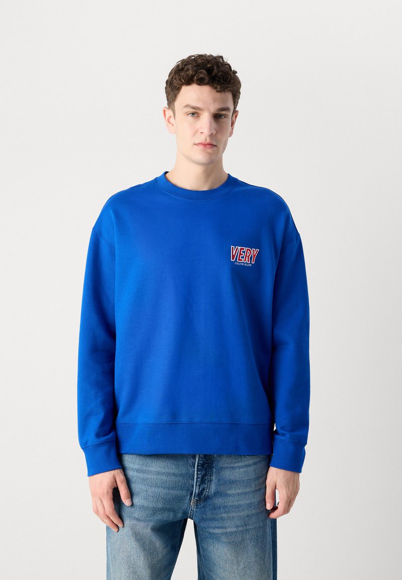 Sudadera azul con cuello redondo acanalado, hombros caídos y un logo frontal que dice "VERY" en rojo y blanco. Combinada con jeans de color azul claro.