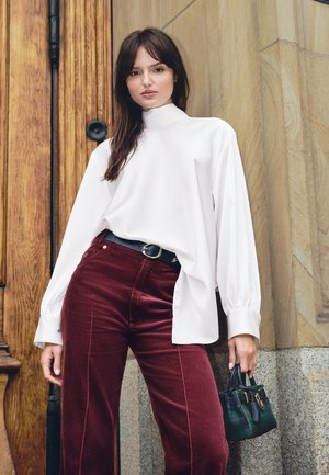 Une femme porte un chemisier blanc à col montant, un pantalon bordeaux et un sac à main noir, près d’une porte en bois et d’un mur en pierre.