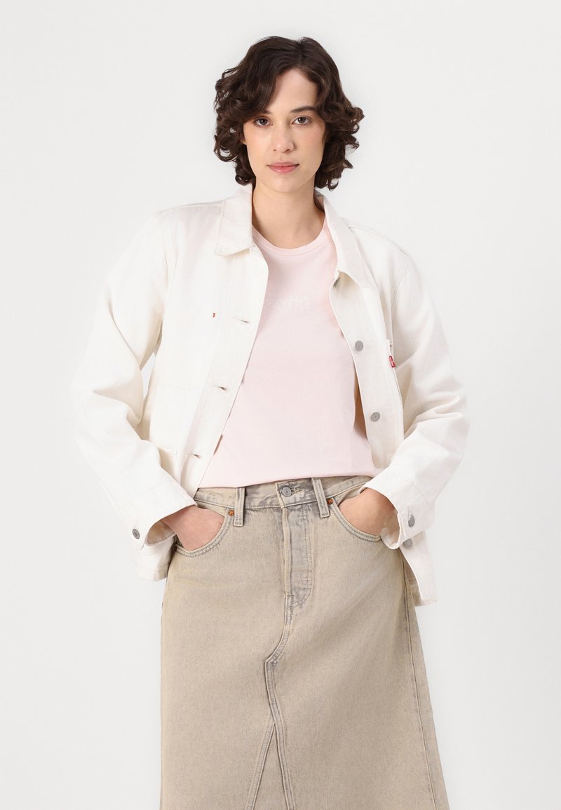 Veste en denim crème avec des boutons argentés sur un t-shirt rose clair, associée à une jupe en denim beige. La mannequin se tient les mains dans les poches.