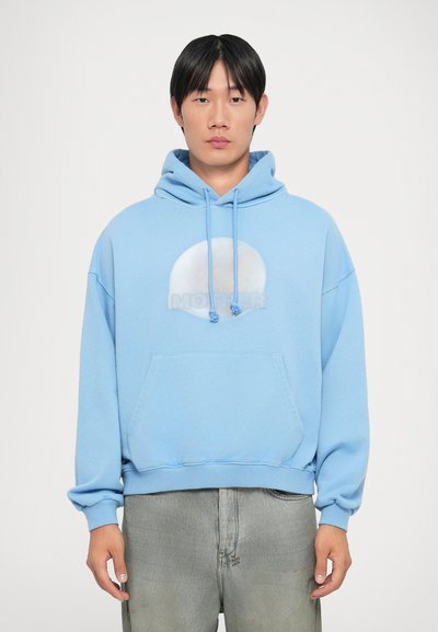 Lichtblauwe hoodie met een voorzak, een capuchon met trekkoord en een ronde grafiek met het woord "MOEDER" in het midden. Zachte textuur.