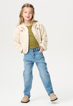 Cremfarbener gesteppter Bomberjacke mit floralem Stickereien, grünes T-Shirt, blaue hochtaillierte Jeans und hellbraune Slipper.