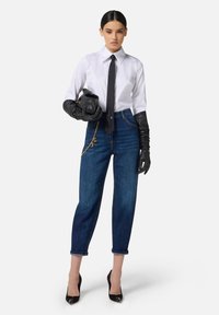 Camicia bianca a maniche lunghe, cravatta nera, jeans a vita alta blu scuro, guanti in pelle nera e tacchi a punta con finitura lucida. Dettaglio in catena dorata.