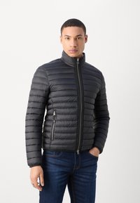 Veste matelassée noire avec un design en losanges, col montant et poches zippées sur les côtés. Portée avec un jean bleu foncé.