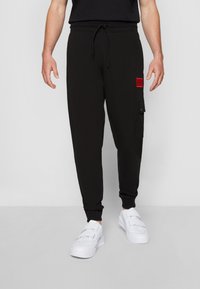 Zwarte joggers van zachte stof, met een trekkoord in de taille, taps toelopende boorden en een rood logo-op een linkerdij. G gedragen met witte sneakers.