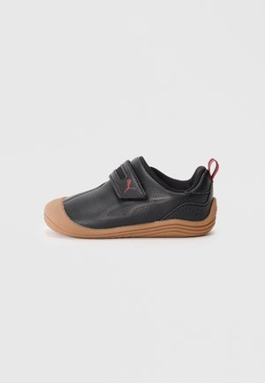 Chaussure de sport noire avec une tige en cuir texturé, une semelle en caoutchouc beige et une sangle en Velcro. Dotée d'un logo accentué en rouge et d'une languette à l'arrière.