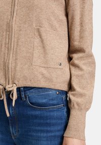 Beige gestrickte Strickjacke mit einem Reißverschluss vorne, einer Tasche, Kordelzug zur Anpassung des Saums und gerippten Bündchen. Getragen über blauen Jeans.