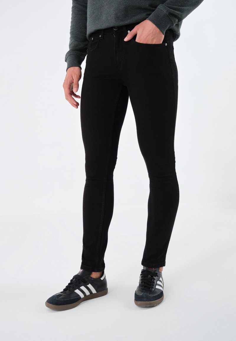 Homme portant un jean slim noir et des baskets Adidas noires avec des bandes blanches, debout avec une main dans la poche.