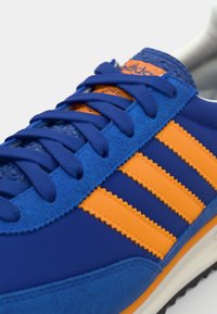 Blue athletic shoe with orange stripes; made of smooth synthetic material and suede; features flat laces and textured accents.