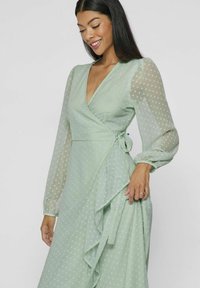 Lys grønn prikkete omslagskjole med gjennomsiktige lange ermer, en dyp V-hals, og en ruffle-detalj på siden. Stoffet ser flytende og teksturert ut.