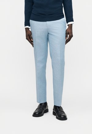 Homme portant un pantalon sur mesure bleu clair associé à des mocassins en cuir noir et un pull bleu foncé avec des poignets de chemise blanche visibles.
