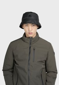 Cappello a secchiello nero con visiera testurizzata e logo impresso; il modello indossa una giacca olive green con zip, colletto alto e tasche.