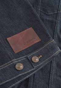 Temno moder denim material z vidno teksturo, s svetlo rjavim usnjenim oznakami z vtisnjenim napisom "GABBA" in kovinskimi gumbi spredaj.