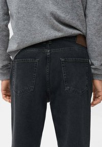 Jeans de mezclilla negra con un ajuste recto, que presentan dos bolsillos traseros y un parche de cuero marrón en la cinturilla. Suéter gris visible arriba.