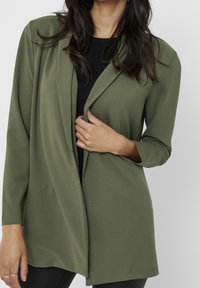 Blazer verde oliva con una tela suave, diseño de frente abierto y mangas tres cuartos. Presenta un drapeado sutil y un cuello de corte relajado.