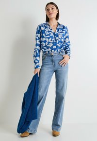 Camisa de manga larga azul con estampado floral y cuello, vaqueros de pierna ancha azul claro y un chal de punto azul oscuro con textura. Botines de tacón color marrón.