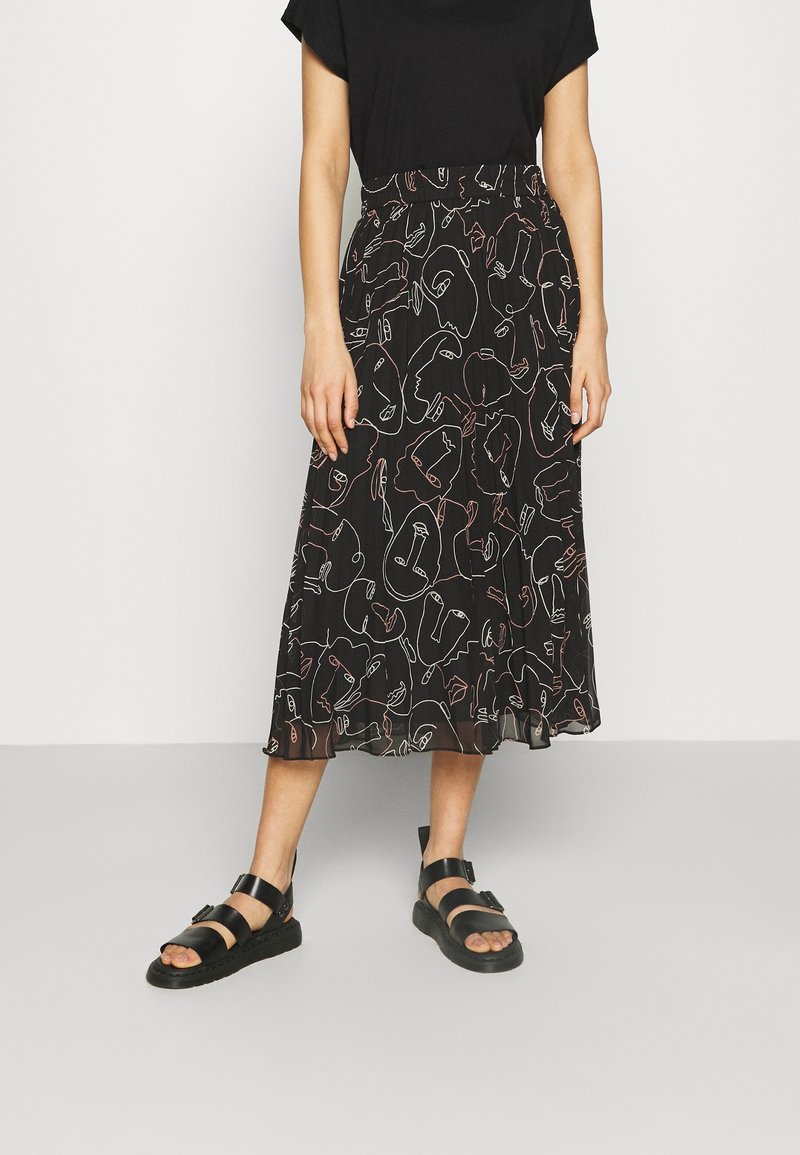 Femme portant une jupe midi noire avec des visages abstraits en art linéaire blanc et rose, un haut noir et des sandales noires avec des boucles.