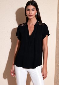Top nero a maniche corte con scollatura a V, dettagli in pizzo sulle spalle e rifinitura decorativa lungo la parte anteriore. Abbinato a pantaloni bianchi.