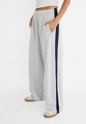 Persoon draagt lichtgrijze sweatpants met brede pijpen en zwarte zijstrepen, handen in de zakken, gecombineerd met witte sneakers en een wit cropped topje.