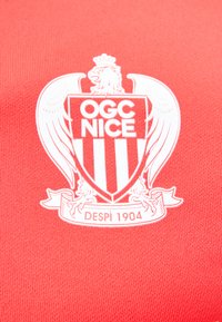 Blason blanc du club de football OGC Nice avec aigle et "Despi 1904" sur fond de tissu rouge.