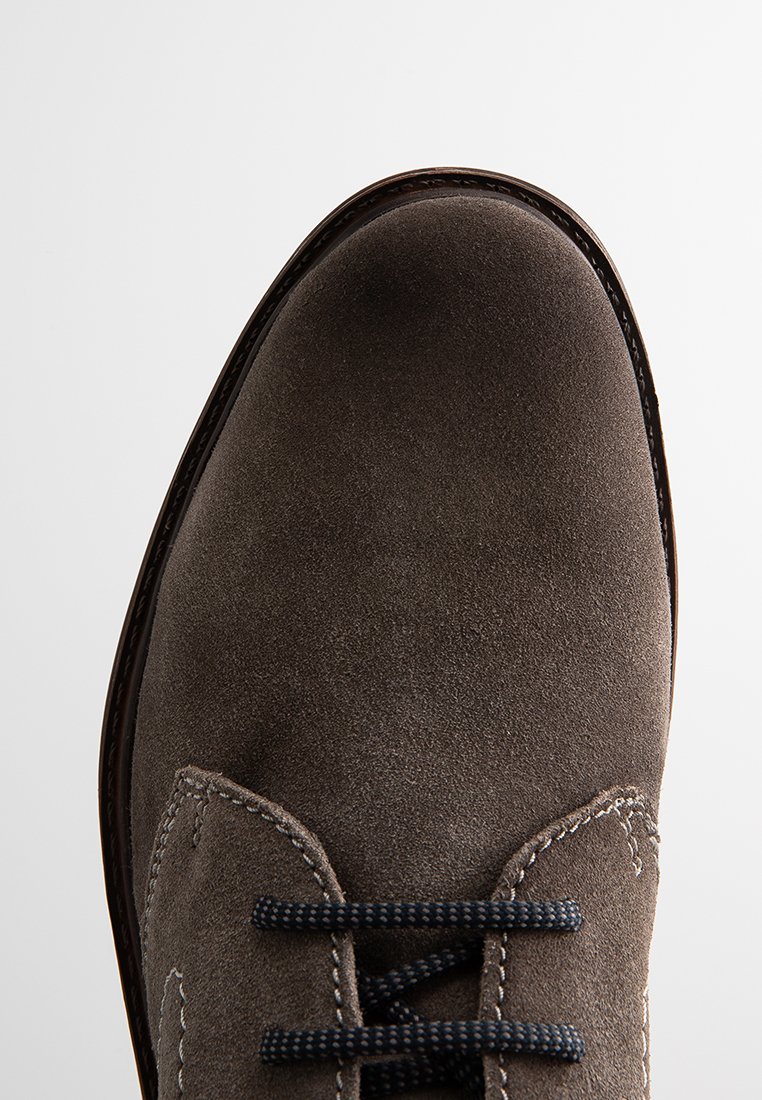 Zapato de gamuza gris con puntera redonda, con cordones azul oscuro y detalles de costura a lo largo del costado, sobre un fondo blanco.