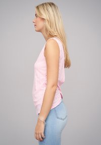 Roze mouwloos topje met oogjespatroon en gebogen zoom, gecombineerd met lichtblauwe slim-fit jeans. Model staat in profiel tegen een grijze achtergrond.