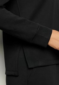 Primo piano di una persona che indossa una maglietta nera a maniche lunghe e pantaloni neri, con un evidente spacco laterale e dettagli di cucitura all'orlo.
