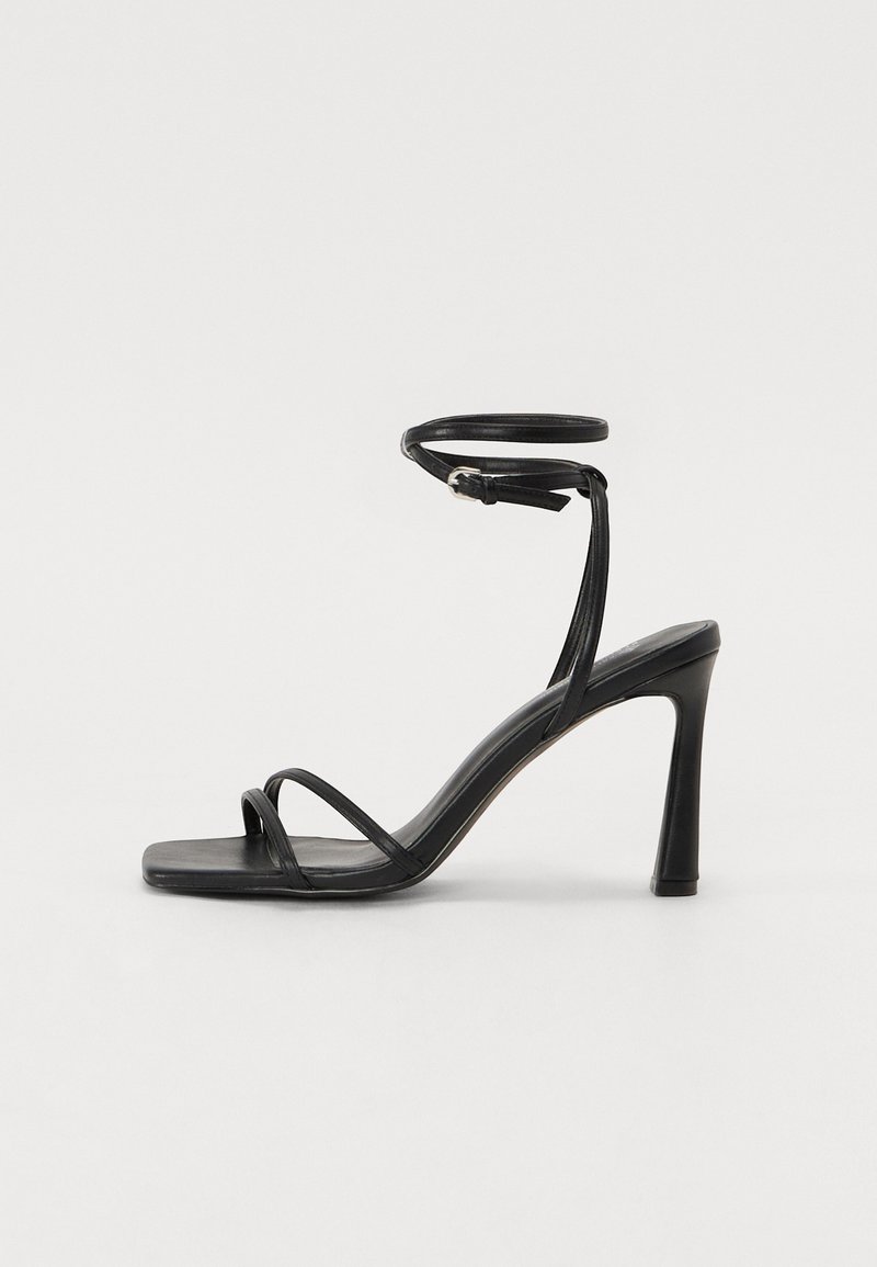Sandal nera in pelle con tacco alto, dotata di sottile cinturino alla caviglia, design a punta quadrata e multiple fasce strette che attraversano il piede. Texture liscia.