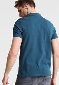 Türkisfarbenes Polo-Shirt mit kurzen Ärmeln, weicher, glatter Textur und umgeschlagenem Kragen. Es hat einen geraden Saum und dezente Nähte auf der Rückseite.