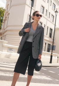 Hall hall villane bleiser halli nööbitava topi peal, mustade kulottidega ning hoides käes kootud musta käekotti. Ülepaisutatud päikeseprillid täiustavad välimust.