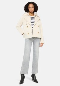 Beige zweireihige Jacke mit Kapuze, goldfarbenen Knöpfen und zwei Taschen, kombiniert mit gestreiftem Hemd und hellgrauen Schlagjeans. Schwarze Stiefeletten.