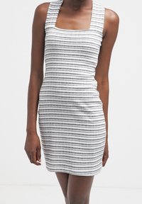 Vrouw in een mouwloos, knielang wit jurkje met dunne horizontale zwarte strepen, staand tegen een effen witte achtergrond.