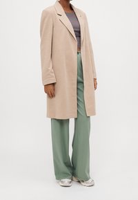 Persona wearing un abrigo largo beige, un top corto gris, pantalones verdes holgados y zapatillas blancas, de pie contra un fondo claro y liso.