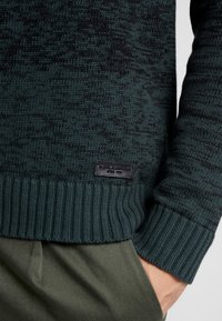 Pull en maille vert foncé avec un motif texturé, des poignets côtelés et une étiquette en tissu noir sur le côté inférieur. Associé à un pantalon olive.
