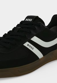 Baskets en cuir noir avec bandes blanches sur les côtés et semelle marron, avec marquage "BOSS" sur la languette et le côté, lacets noirs détaillés.