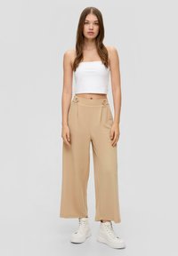 Beige wijde broek met plooien en knoopaccenten in de taille, gecombineerd met een cropped wit tanktop en witte hoge sneakers.