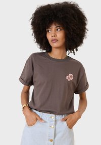 Camiseta de algodón marrón con bordado floral en rosa en la parte izquierda del pecho, mangas cortas enrolladas, combinada con una falda de mezclilla clara.