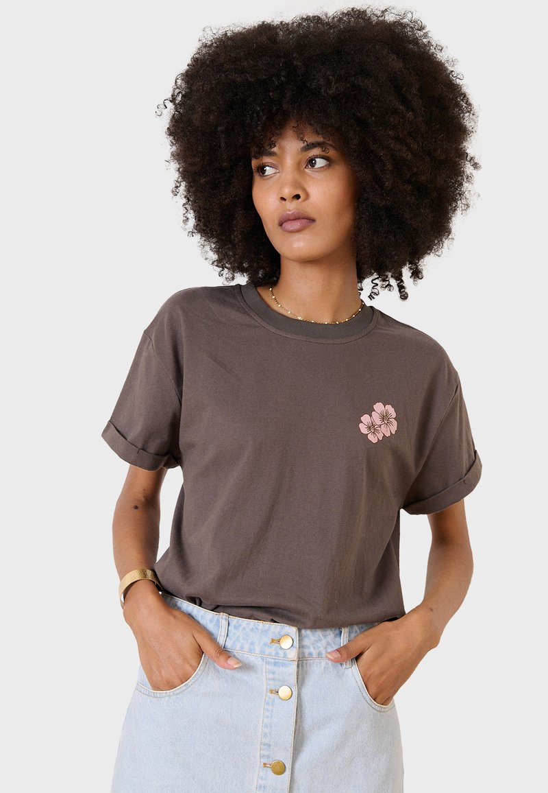 Camiseta de algodón marrón con bordado floral en rosa en la parte izquierda del pecho, mangas cortas enrolladas, combinada con una falda de mezclilla clara.