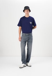 Marineblauwe katoenen t-shirt met een logo, lichtblauwe denim jeans, grijs-witte sneakers en een zwarte bucket hat, staande tegen een witte achtergrond.