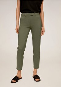 Pantalon vert olive à coupe slim, texture lisse, avec poches avant ; porté avec un haut noir et des sandales plates noires.