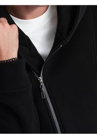 Černá mikina na zip s hladkým povrchem, se stříbrným zipem a bílým tričkem pod ní. Manžetové rukávy s uvolněným střihem.