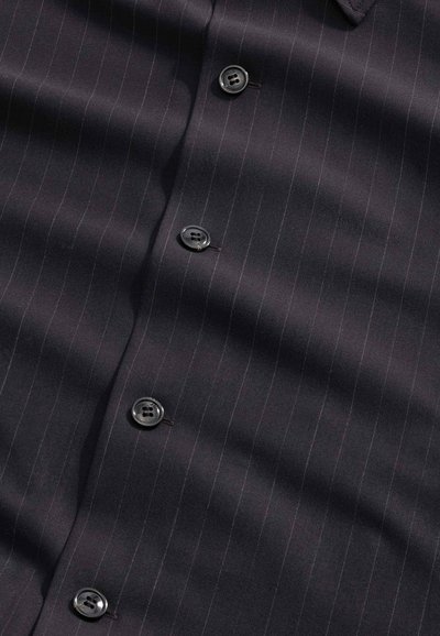Tissu à fines rayures sombre avec quatre boutons noirs cousus verticalement au centre, montrant une texture subtile et des plis.