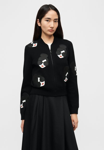 Crni džemper na patentni zatvarač s ponavljajućim vezanim portretima s naočalama i crvenim usnama. Uže manžetne i rub, mekana tekstura.